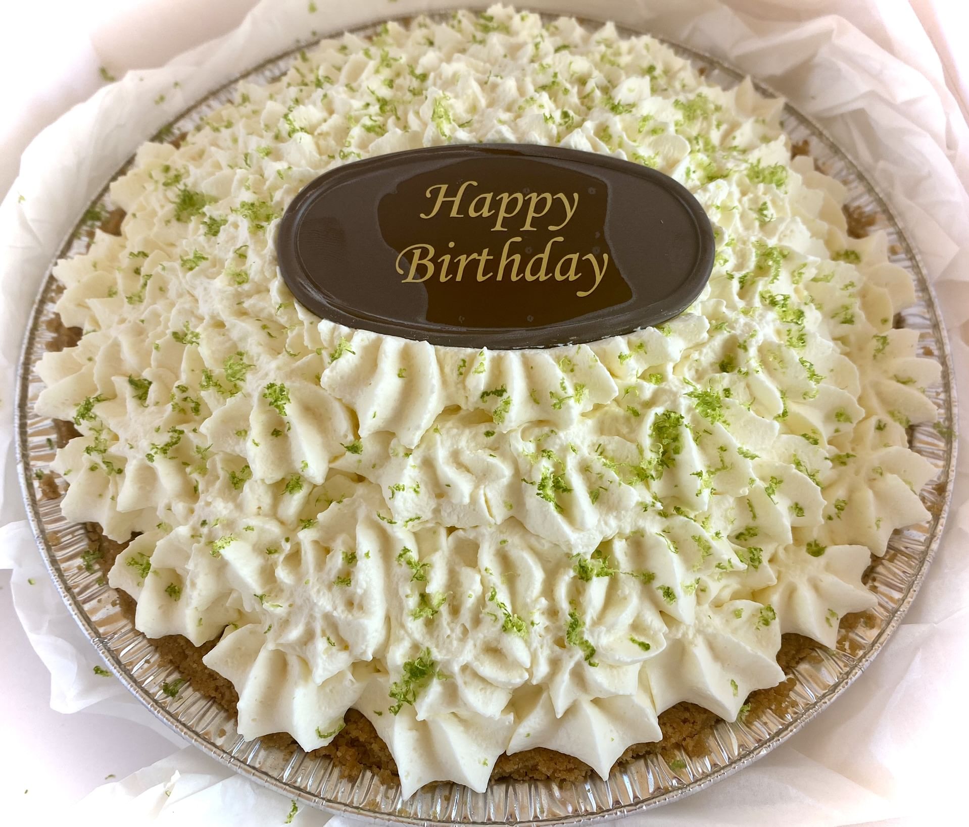 A decorative birthday Key Lime Pie topped with a layer of swirled cream and sprinkled with green zest. A dark oval chocolate plaque in the center reads 'Happy Birthday' in golden letters. The pie, made with authentic Key West key limes, is presented in a tin pie dish.